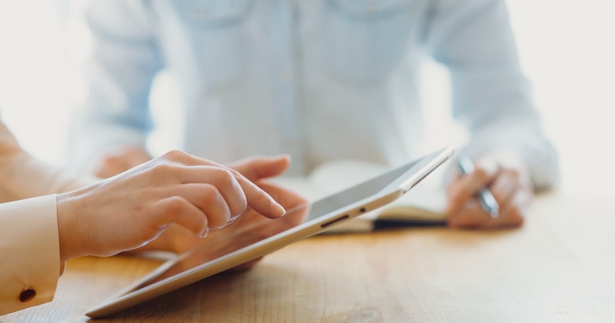 A person working on an iPad at a wooden table and another in the background writing in a book.