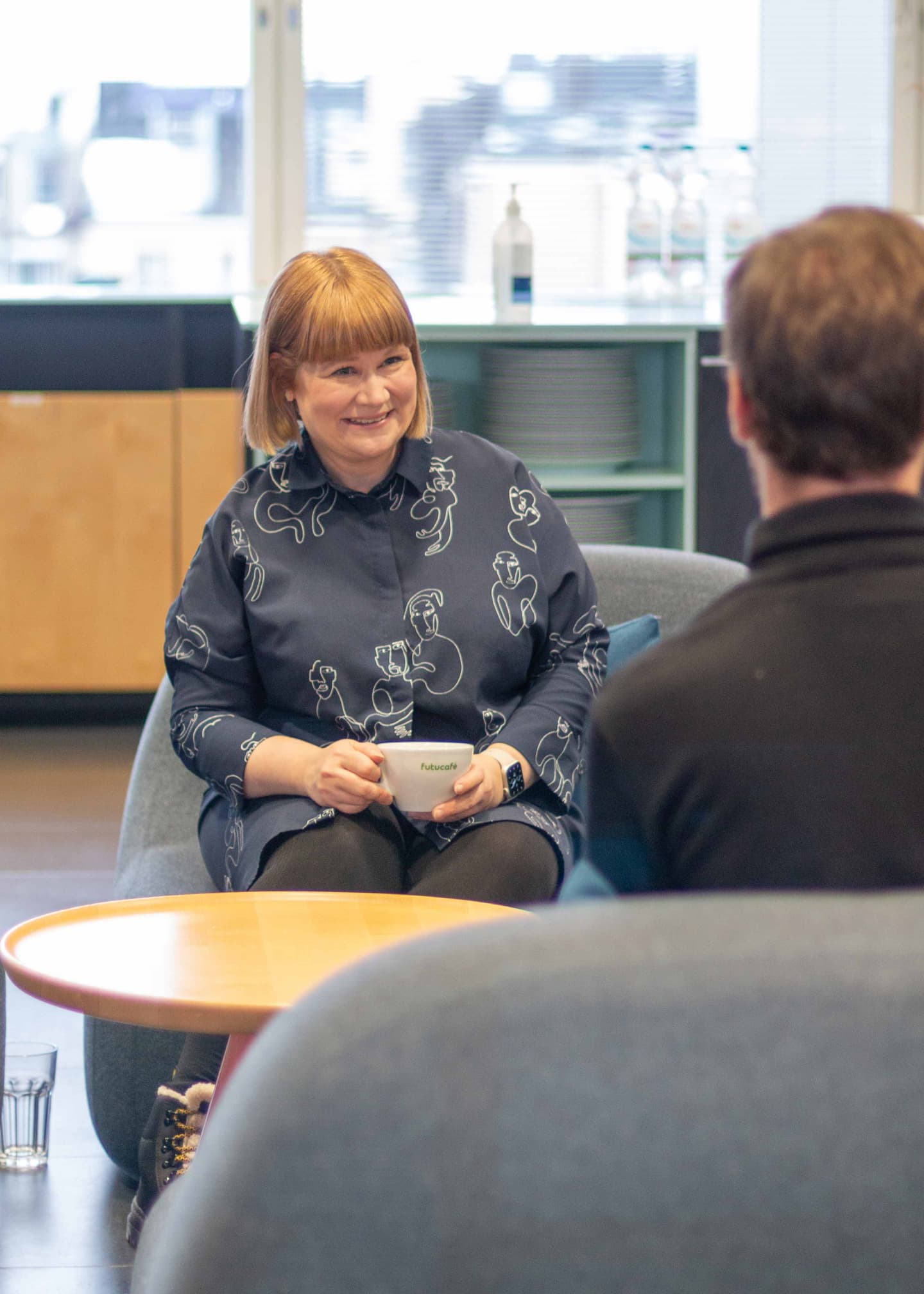 Person in blue patterned shirt holding a coffee cup during conversation in modern office with city view.