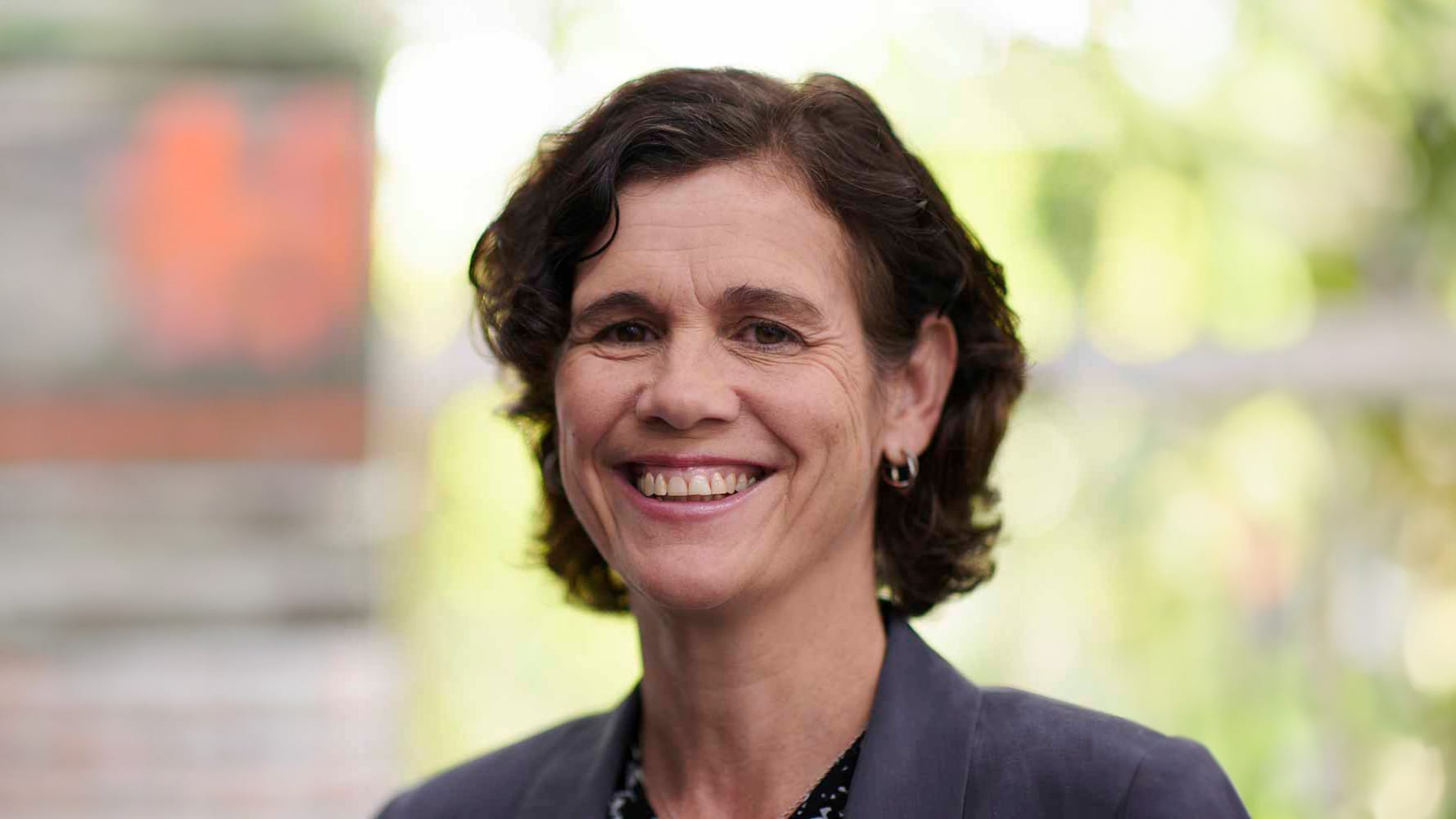 Person with short wavy dark hair smiling broadly, wearing a dark blazer against a soft-focus green background.