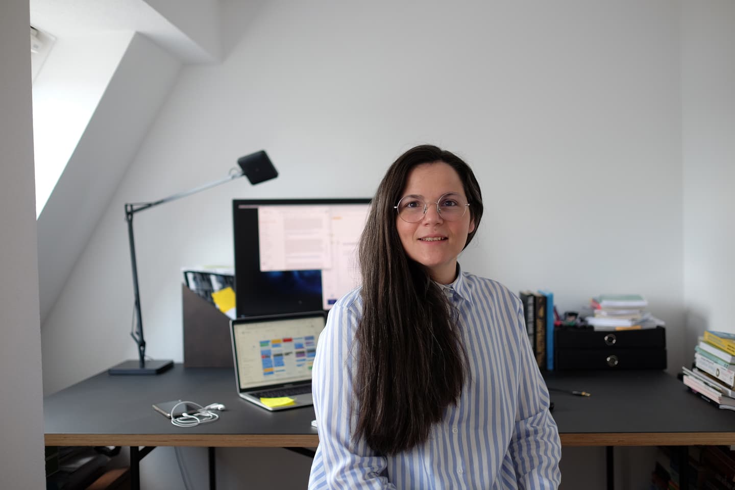 Person with long dark hair and glasses in a blue striped shirt working at a home office desk with computer monitors.