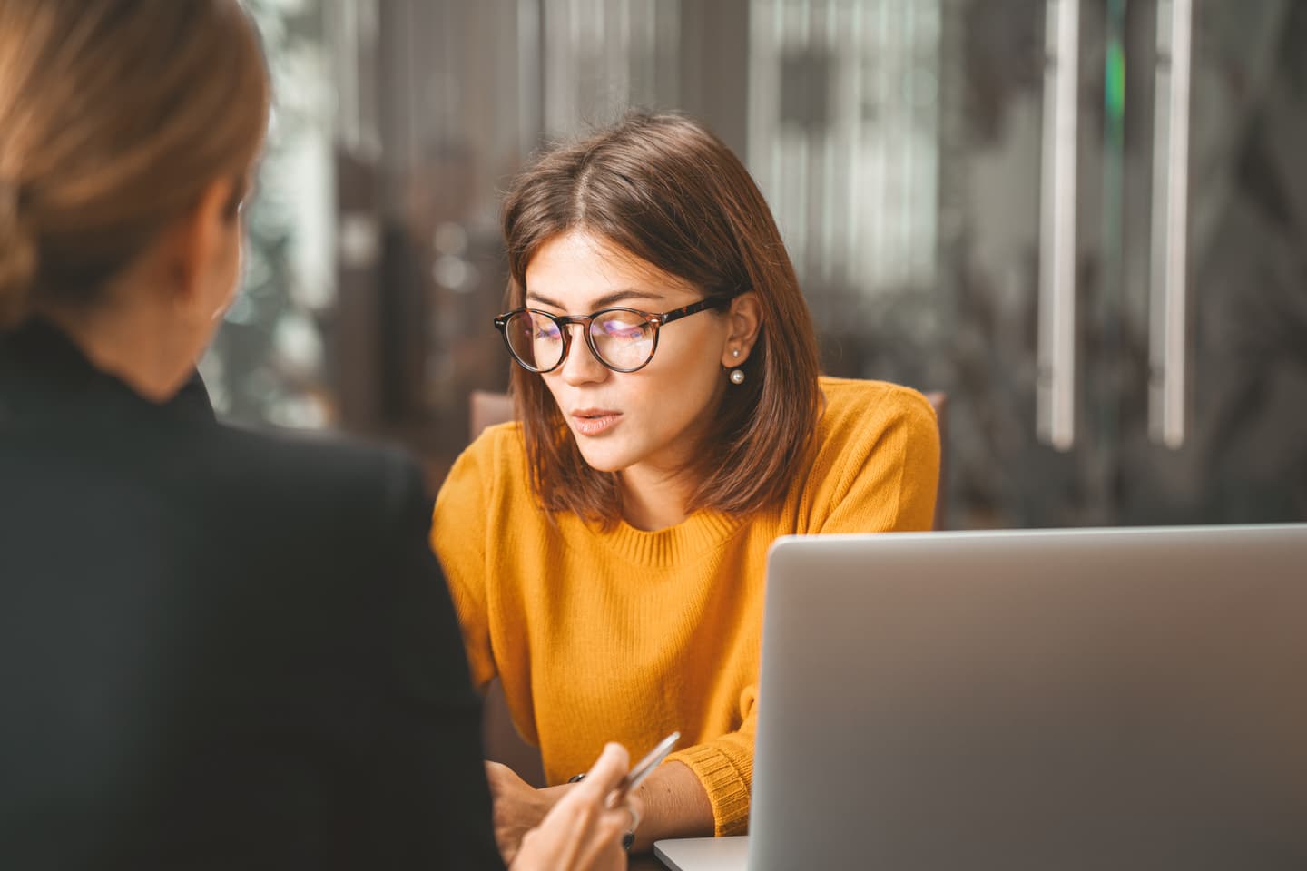Person in bright yellow jumper wearing glasses focused on laptop during a business meeting in modern office setting.