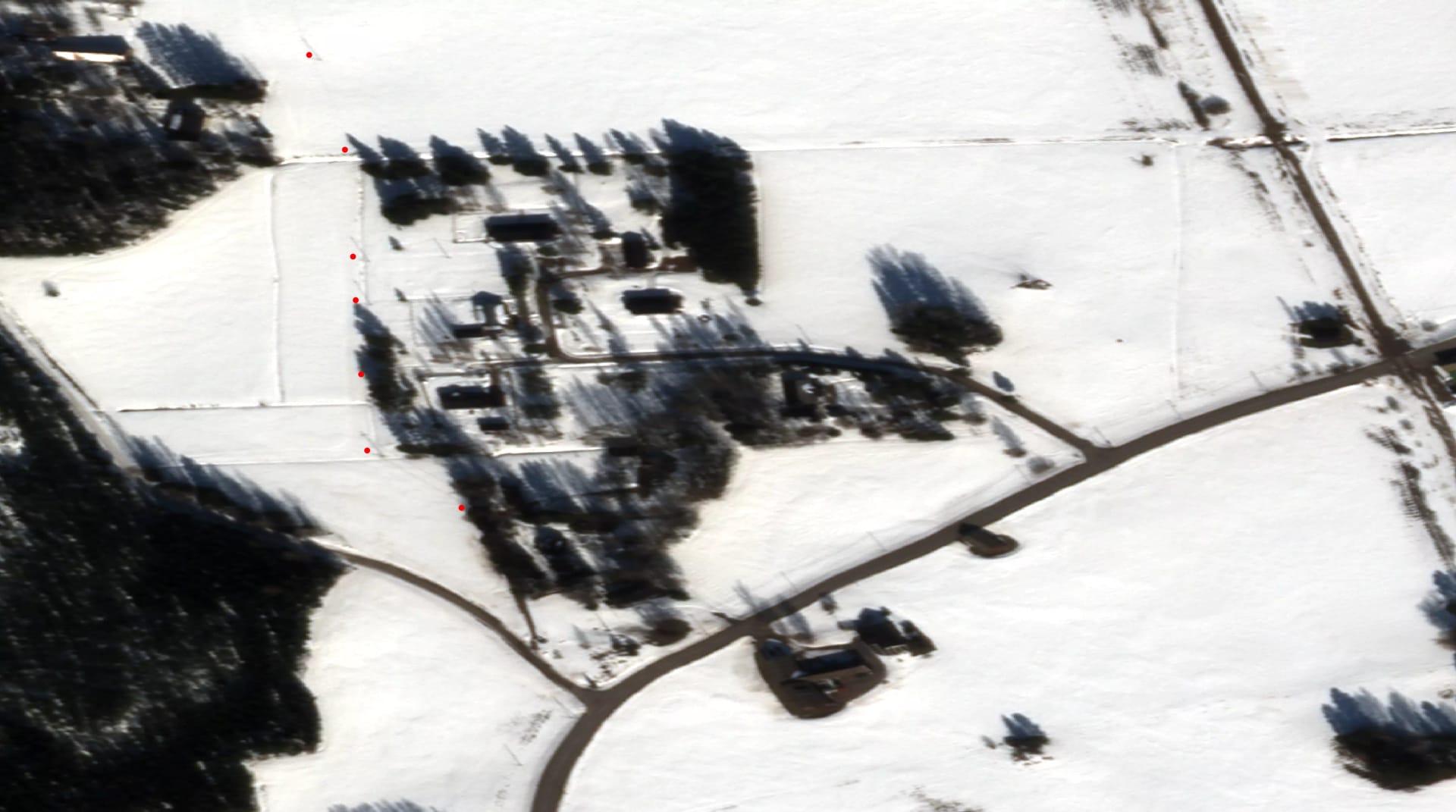 Aerial view of a rural property in winter, showing snow-covered fields, buildings, and roads with dark trees scattered throughout.
