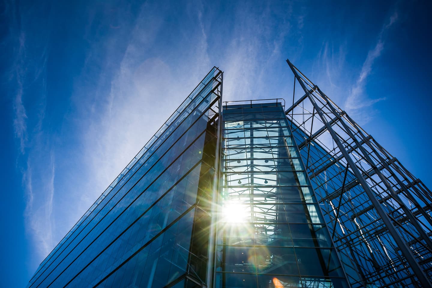 Modern glass skyscraper viewed from below with sunlight shining through its transparent structure against a blue sky.