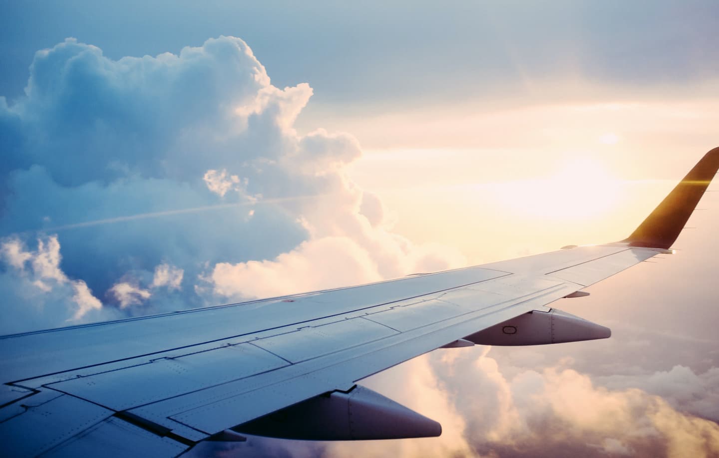 Aeroplane wing against a sunset sky with fluffy white clouds, sunlight creating a golden glow on the horizon.