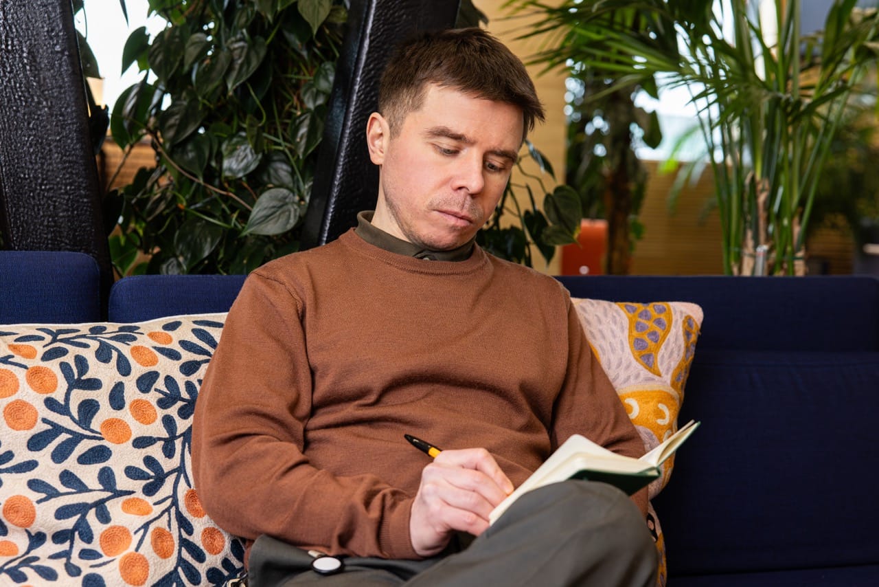 A man in a brown jumper sits on a navy sofa writing in a notebook, surrounded by colourful cushions and green plants.