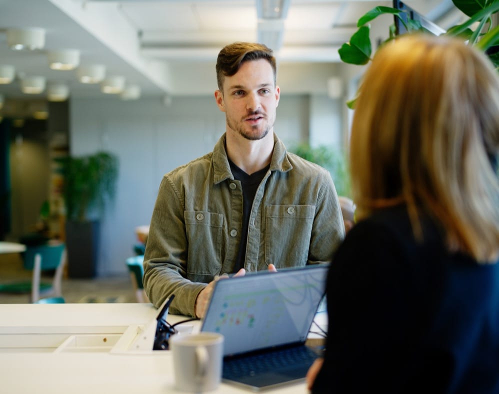 Two people in conversation at a modern office with plants, laptop open on table and coffee cup nearby.