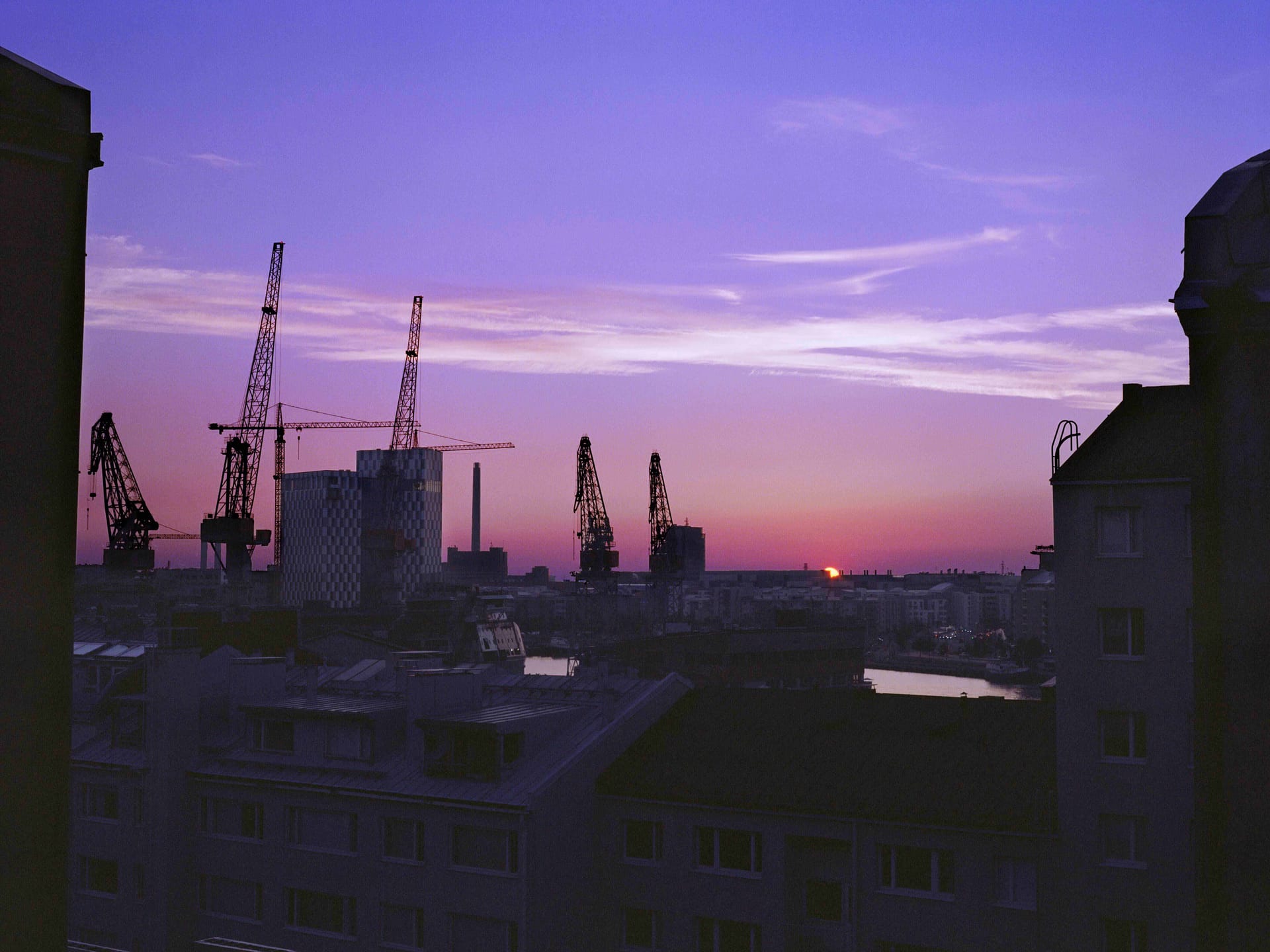 Urban skyline at sunset with construction cranes silhouetted against a vibrant purple sky, buildings in foreground.