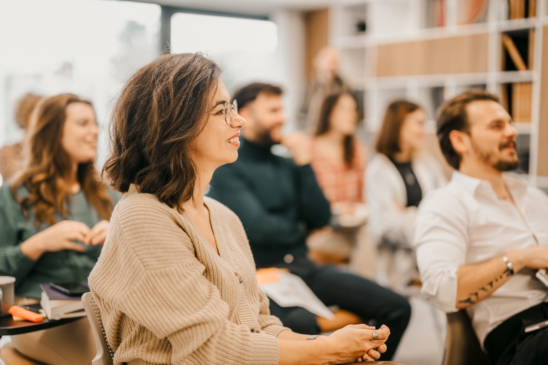 Person in beige cardigan with glasses smiling during a presentation, with audience members in background.