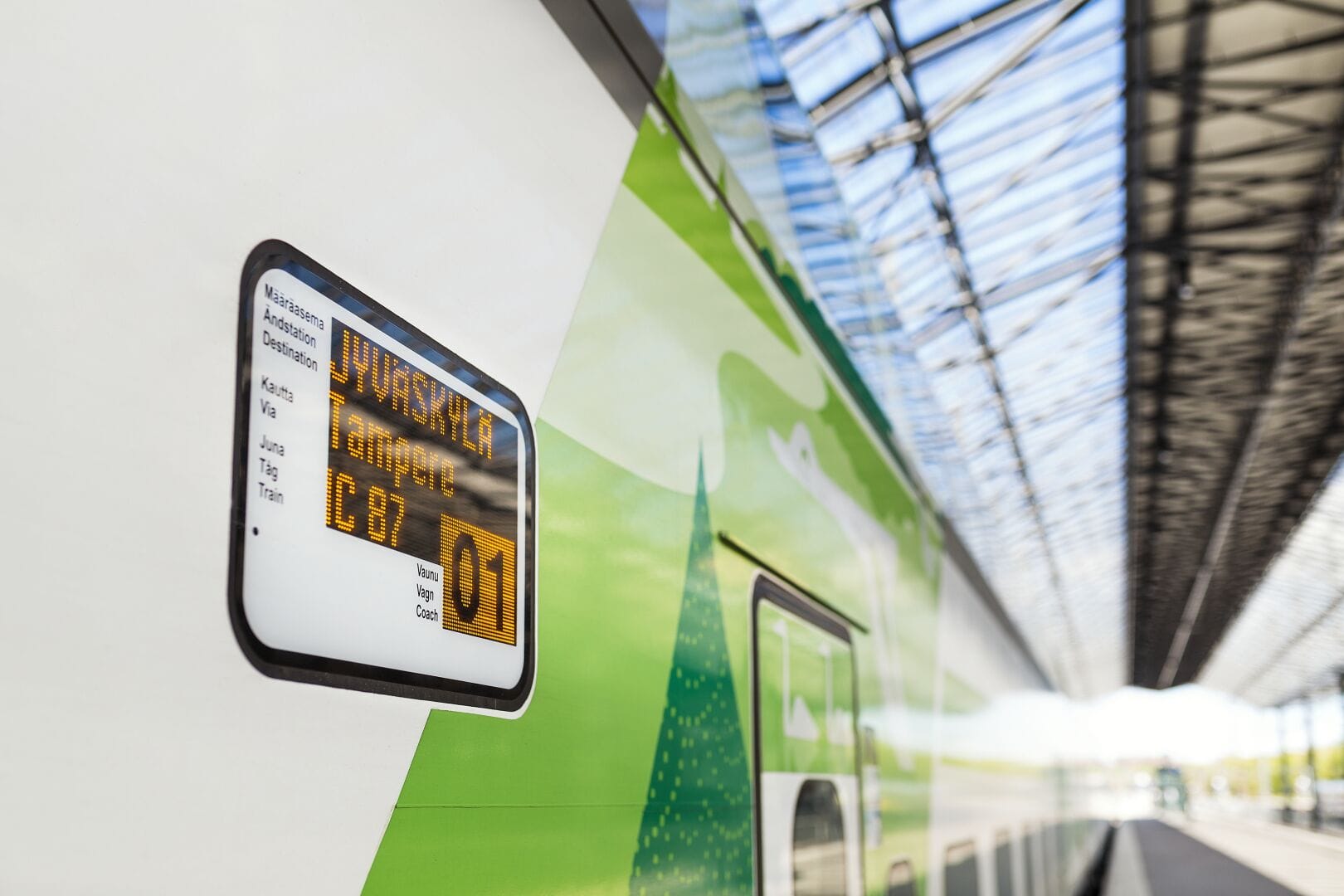 Digital train display showing destination Jyväskylä on a green and white train at a station platform under glass roof.