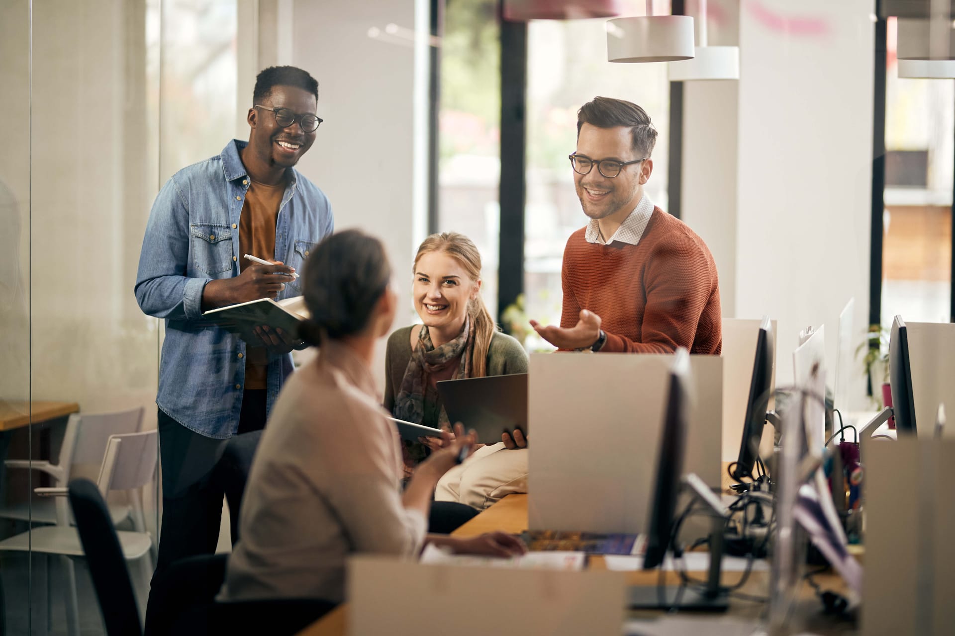 Diverse colleagues having a friendly discussion in a bright modern office space with large windows and computer workstations.