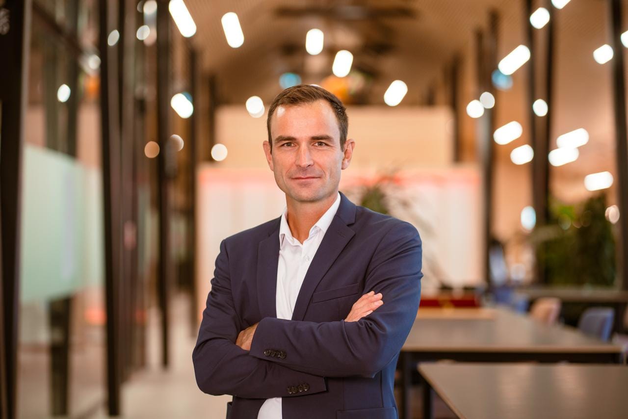 Professional in navy suit with arms crossed standing in modern office corridor with bokeh lighting in background.