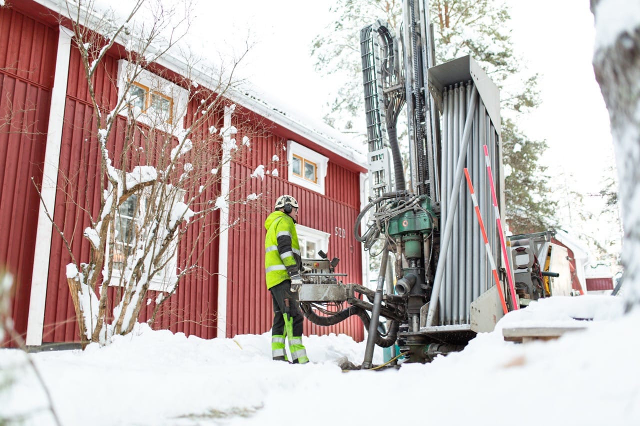 A worker in a hi-vis jacket operates a ground drilling rig in snow beside a red building during winter.