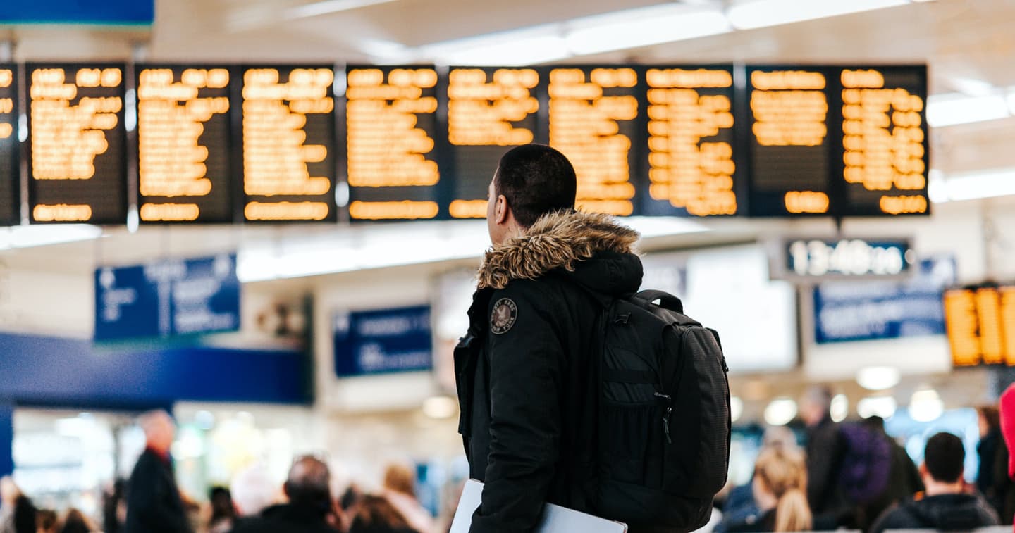 Traveller in winter coat with backpack looking at orange departure boards in busy train station terminal.