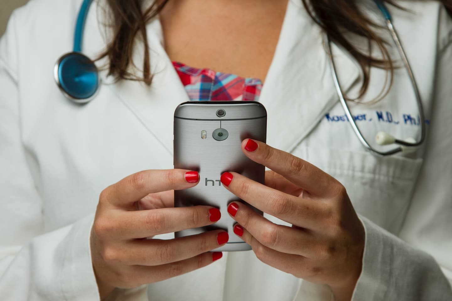 Medical professional in white coat with stethoscope using silver HTC smartphone, displaying red nail polish.