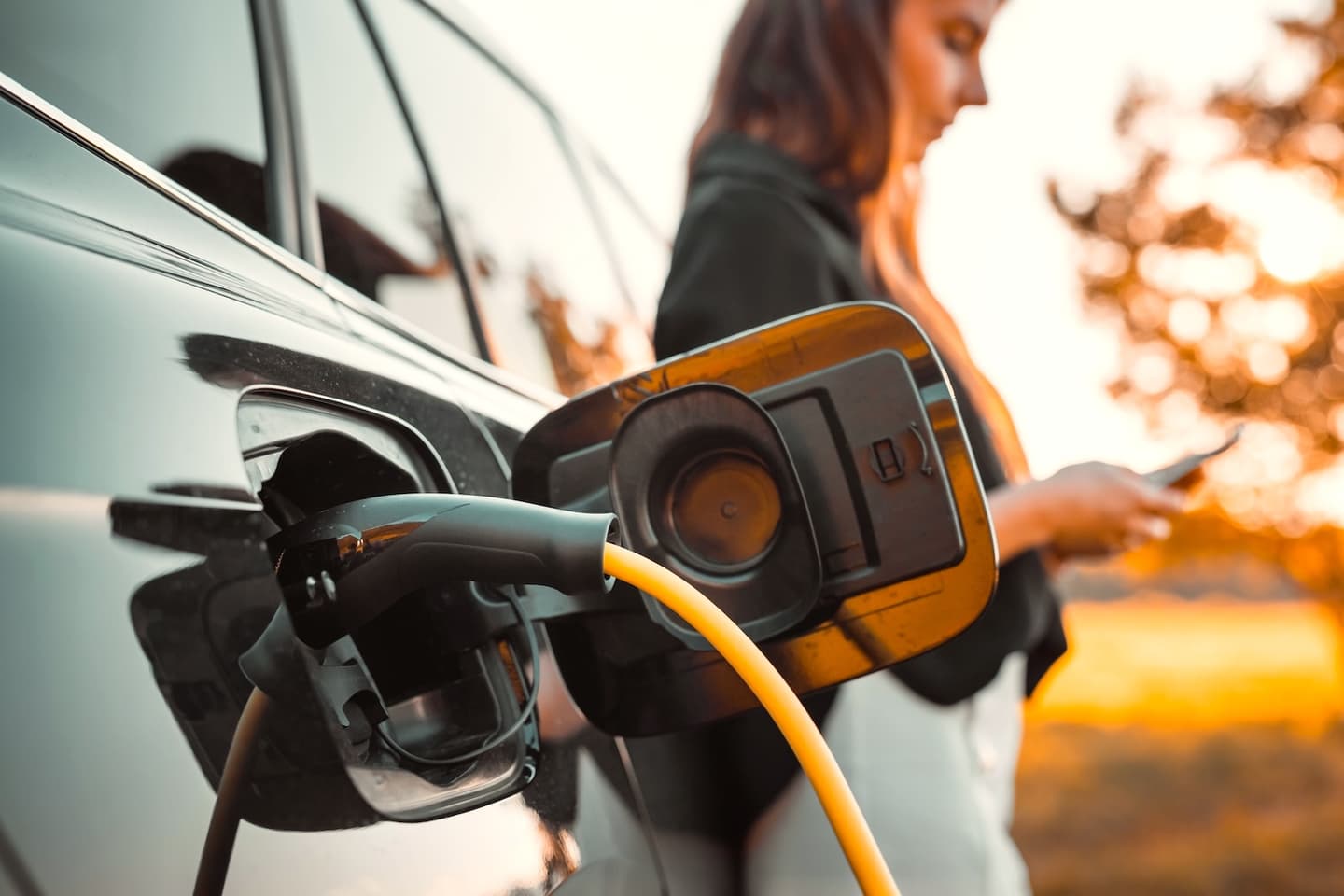 Electric car charging port with yellow cable connected, person standing nearby using their smartphone at sunset.