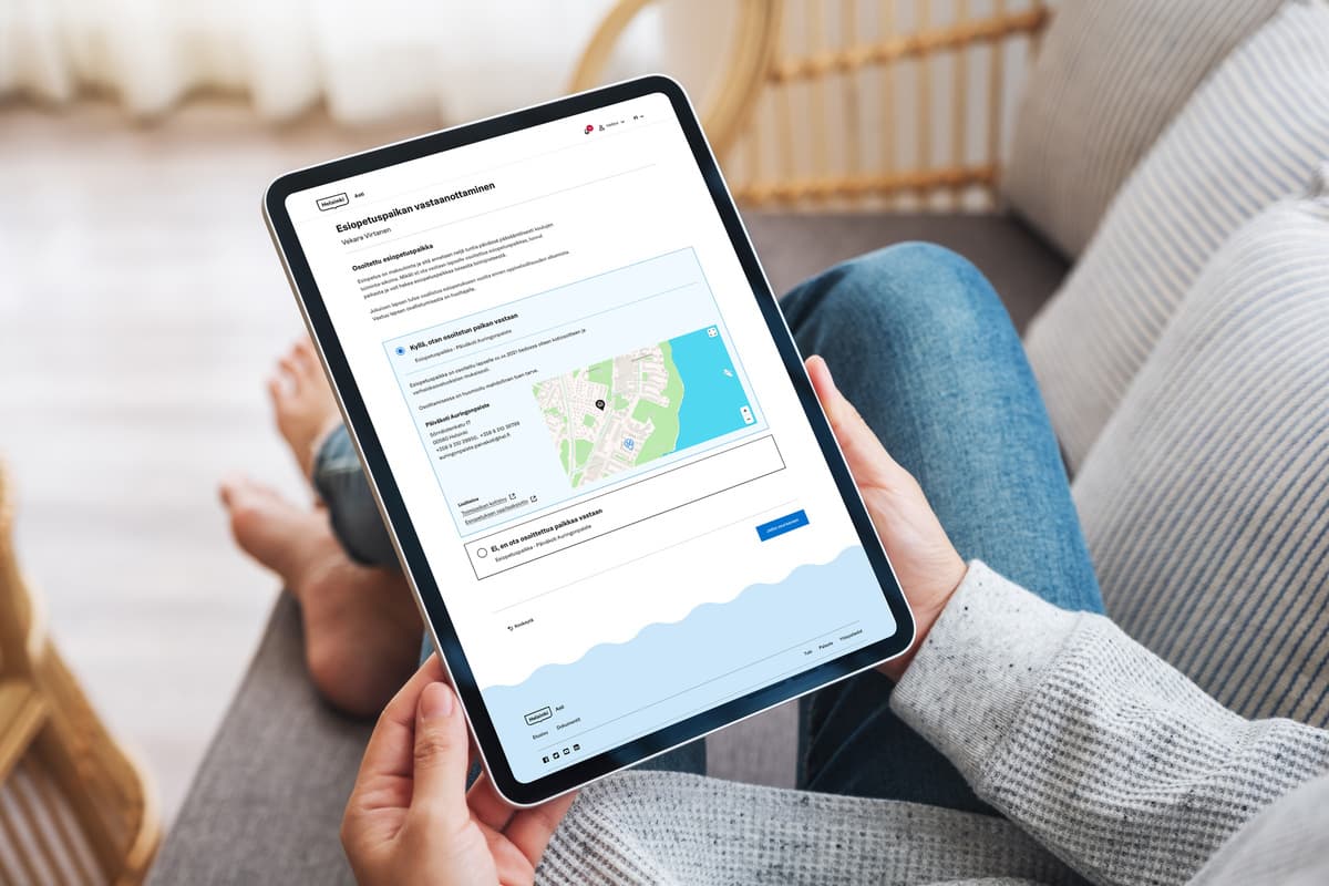 Person in grey jumper using tablet showing Finnish website with map interface, sitting on couch in casual home setting.