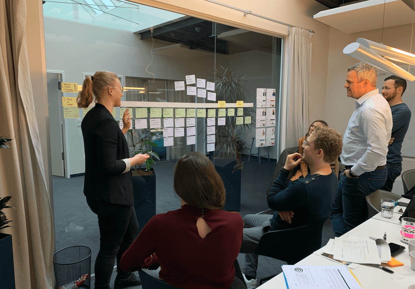 Team meeting with people discussing project plans displayed on glass wall with colourful sticky notes and documents.